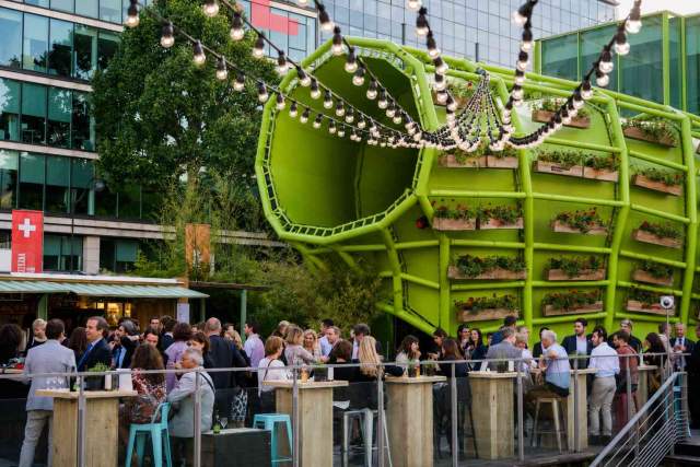 A lively House of Switzerland outdoor event space, with guests gathering around wooden tables beneath string lights and a distinctive green installation.