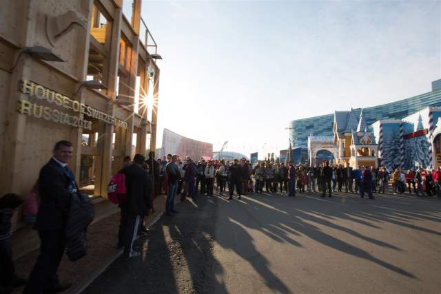 A crowd gathered outside the House of Switzerland Russia 2014, with the wooden Swiss pavilion illuminated by the low afternoon sun. Visitors line the street alongside other national pavilions, creating a lively event atmosphere.