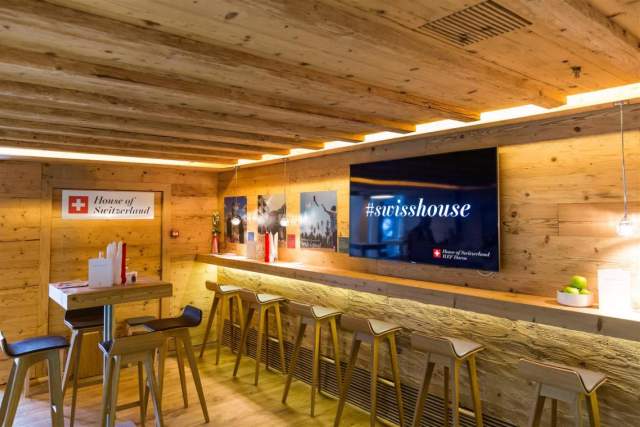 An interior view of the House of Switzerland, featuring a warm wooden lounge with bar seating, Swiss branding and a large screen displaying the hashtag #swisshouse.